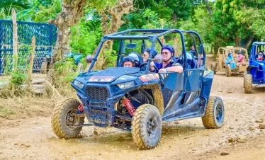 Four people wearing helmets ride in a blue off-road buggy on a dirt path surrounded by greenery during one of the exciting Punta Cana excursions. Another buggy follows, as the muddy tires show their recent adventure through rugged terrain.