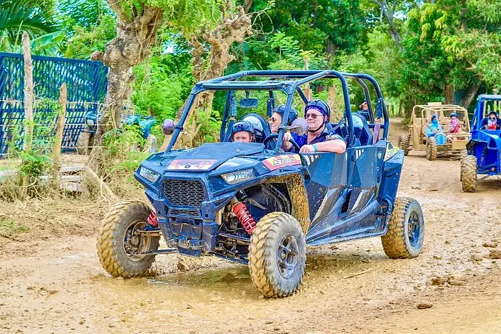 Four people wearing helmets ride in a blue off-road buggy on a dirt path surrounded by greenery during one of the exciting Punta Cana excursions. Another buggy follows, as the muddy tires show their recent adventure through rugged terrain.