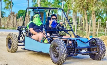 Two people in sunglasses and face coverings ride a blue dune buggy with large off-road tires on a dirt path. Surrounded by green trees and wooden fences, they give a thumbs-up—capturing the thrill of Punta Cana tours in action.