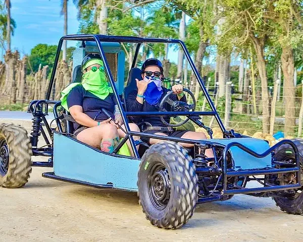 Two people in sunglasses and face coverings ride a blue dune buggy with large off-road tires on a dirt path. Surrounded by green trees and wooden fences, they give a thumbs-up—capturing the thrill of Punta Cana tours in action.
