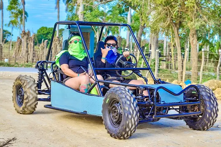 Two people in sunglasses and face coverings ride a blue dune buggy with large off-road tires on a dirt path. Surrounded by green trees and wooden fences, they give a thumbs-up—capturing the thrill of Punta Cana tours in action.