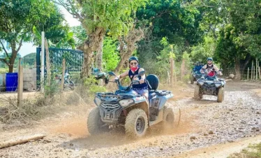 Two people ride ATVs on a dirt path lined with trees and vegetation during a sunny Punta Cana trip. The lead rider, wearing sunglasses and a helmet, smiles while splashing through a muddy puddle as the second follows closely behind.