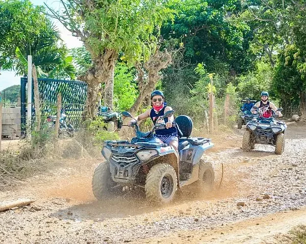 Two people ride ATVs on a dirt path lined with trees and vegetation during a sunny Punta Cana trip. The lead rider, wearing sunglasses and a helmet, smiles while splashing through a muddy puddle as the second follows closely behind.