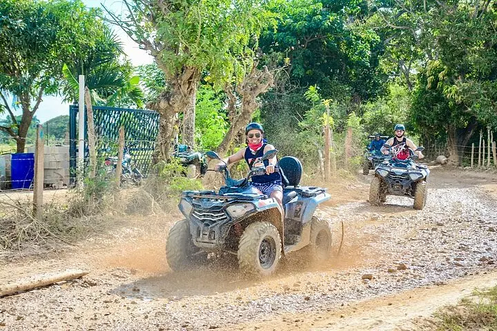 Two people ride ATVs on a dirt path lined with trees and vegetation during a sunny Punta Cana trip. The lead rider, wearing sunglasses and a helmet, smiles while splashing through a muddy puddle as the second follows closely behind.