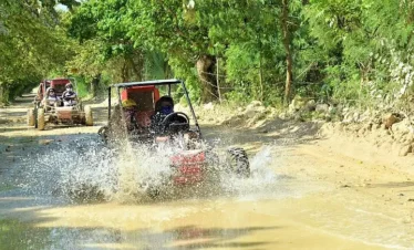 Two off-road buggies drive along a dirt path, splashing through mud as part of exciting Punta Cana excursions. Surrounded by lush trees and sunlit foliage, both vehicles carry helmeted riders enjoying unforgettable Punta Cana tours.