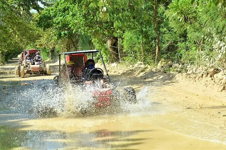 Two off-road buggies drive along a dirt path, splashing through mud as part of exciting Punta Cana excursions. Surrounded by lush trees and sunlit foliage, both vehicles carry helmeted riders enjoying unforgettable Punta Cana tours.