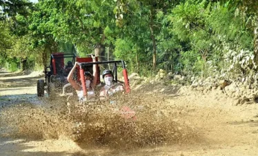 Two people in a dune buggy drive through a large muddy puddle on a dirt road, creating a big splash during one of the exciting Punta Cana excursions. Another buggy follows behind, surrounded by lush greenery, as the drivers wear helmets and sunglasses.