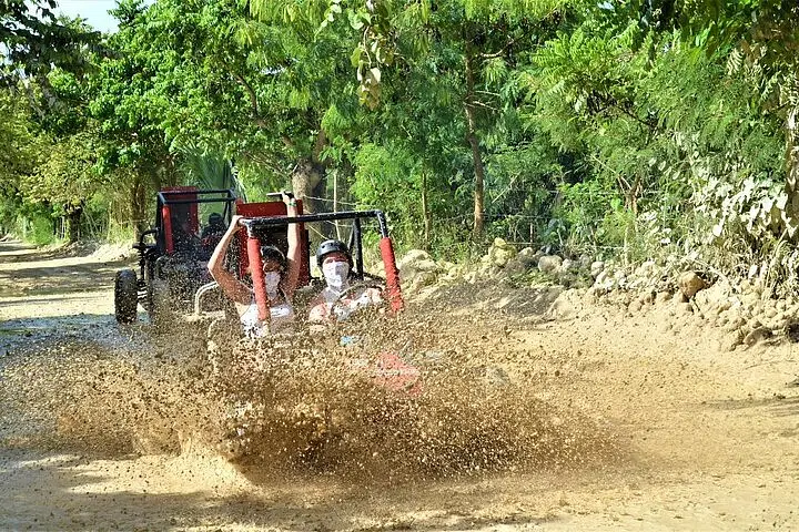 Two people in a dune buggy drive through a large muddy puddle on a dirt road, creating a big splash during one of the exciting Punta Cana excursions. Another buggy follows behind, surrounded by lush greenery, as the drivers wear helmets and sunglasses.