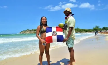 Two people stand barefoot on a sandy beach during their Punta Cana trip, holding a Dominican Republic flag. The woman wears a bikini and glasses; the man wears a hat, sunglasses, and shorts, holding a coconut. Ocean waves and greenery complete the scene.
