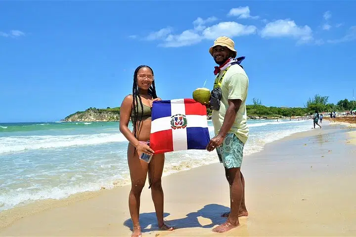 Two people stand barefoot on a sandy beach during their Punta Cana trip, holding a Dominican Republic flag. The woman wears a bikini and glasses; the man wears a hat, sunglasses, and shorts, holding a coconut. Ocean waves and greenery complete the scene.