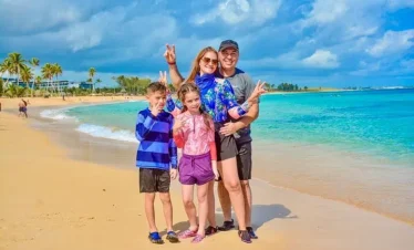 A smiling family of four stands on a sandy beach near turquoise water, enjoying one of their Punta Cana excursions. The parents flash peace signs as their kids pose in front, with palm trees and a blue sky as the perfect backdrop for their adventure.
