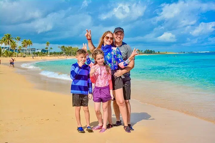 A smiling family of four stands on a sandy beach near turquoise water, enjoying one of their Punta Cana excursions. The parents flash peace signs as their kids pose in front, with palm trees and a blue sky as the perfect backdrop for their adventure.