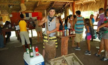 Experience authentic culture on your Punta Cana trip as a man demonstrates traditional techniques in a rustic open-air hut. Guests gather around, enjoying local drinks and condiments during unforgettable tours in Punta Cana.