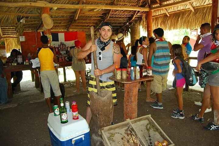 Experience authentic culture on your Punta Cana trip as a man demonstrates traditional techniques in a rustic open-air hut. Guests gather around, enjoying local drinks and condiments during unforgettable tours in Punta Cana.