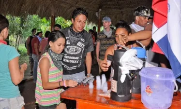 A group of people, including children and adults, gather under a thatched-roof shelter during their Punta Cana tour. A woman pours drinks into small plastic cups on an orange table as others, some in bandanas, wait in line. Tropical plants flourish in the background.