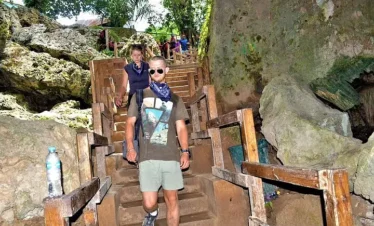 A man in sunglasses, a graphic t-shirt, and a blue scarf walks down rustic wooden stairs in a rocky outdoor area. This scenic spot, perfect for Punta Cana excursions, features lush greenery and large rocks surrounding the path.