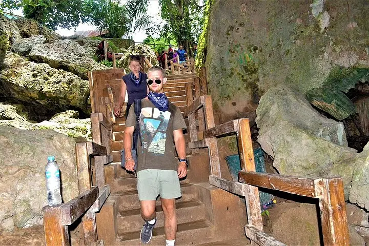 A man in sunglasses, a graphic t-shirt, and a blue scarf walks down rustic wooden stairs in a rocky outdoor area. This scenic spot, perfect for Punta Cana excursions, features lush greenery and large rocks surrounding the path.
