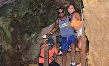 Three people enjoy a cave adventure on their Punta Cana trip. A man in sunglasses and a tank top smiles as a woman in shorts hugs him, while another in a red top and cap leans on the wooden railing, surrounded by rocky cave walls.