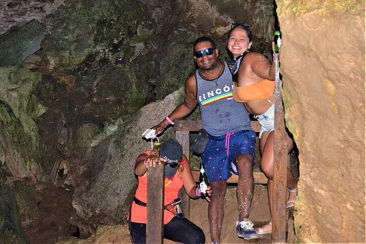Three people enjoy a cave adventure on their Punta Cana trip. A man in sunglasses and a tank top smiles as a woman in shorts hugs him, while another in a red top and cap leans on the wooden railing, surrounded by rocky cave walls.