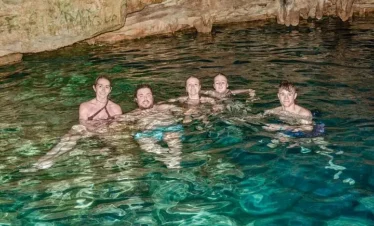 Five people enjoy swimming in a cave pool with clear, turquoise water during one of the top Punta Cana excursions. Smiling at the camera, they’re surrounded by rocky cave walls and ceiling, beautifully lit by artificial light.
