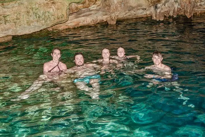 Five people enjoy swimming in a cave pool with clear, turquoise water during one of the top Punta Cana excursions. Smiling at the camera, they’re surrounded by rocky cave walls and ceiling, beautifully lit by artificial light.
