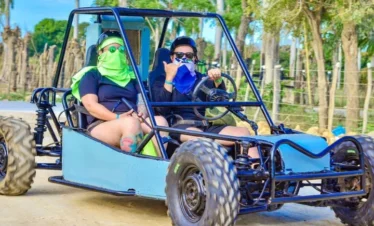 Two people ride in a blue dune buggy on a dirt road during one of the exciting Punta Cana excursions. Both wear sunglasses and bandanas, with the driver making a shaka hand sign. Trees and wooden posts line the background under a bright, blue sky.
