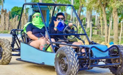 Two people ride in a blue dune buggy on a dirt road during one of the exciting Punta Cana excursions. Both wear sunglasses and bandanas, with the driver making a shaka hand sign. Trees and wooden posts line the background under a bright, blue sky.