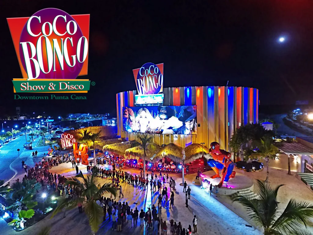 Nighttime aerial view of Coco Bongo Show & Disco in Punta Cana, a must-see on many Punta Cana excursions. The colorful, circular building glows with neon lights as crowds line up outside, palm trees frame the entrance, and a vibrant Coco Bongo sign tops the scene.