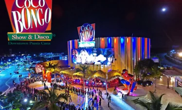 Nighttime aerial view of Coco Bongo Show & Disco in Punta Cana, a must-see on many Punta Cana excursions. The colorful, circular building glows with neon lights as crowds line up outside, palm trees frame the entrance, and a vibrant Coco Bongo sign tops the scene.