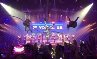 A vibrant stage show with aerial performers, dramatic lights, and a large cast entertains crowds—much like the unforgettable experiences on tours in Punta Cana. The audience holds up phones to record under a bright, illuminated sign.