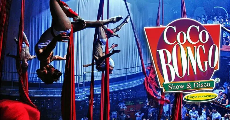 Four aerial silk performers in colorful outfits hang upside down in dramatic poses above a crowded club audience. Bright spotlights highlight the entertainers—a must-see on tours in Punta Cana. A large red and yellow “CoCo Bongo Show & Disco” sign is on the right.