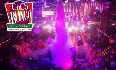 A vibrant nightclub scene at Coco Bongo Punta Cana shows a lively crowd dancing amid colorful lights, smoke, and giant balloons—a must-see for anyone seeking unforgettable Punta Cana tours. The Coco Bongo Show & Disco logo is in the top left corner.