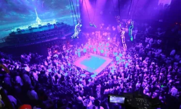 A large crowd surrounds a blue-lit boxing ring in a nightclub with vivid purple lighting—an electrifying scene to experience on your punta cana trip, where performers in glowing suits and digital screens create a futuristic atmosphere.