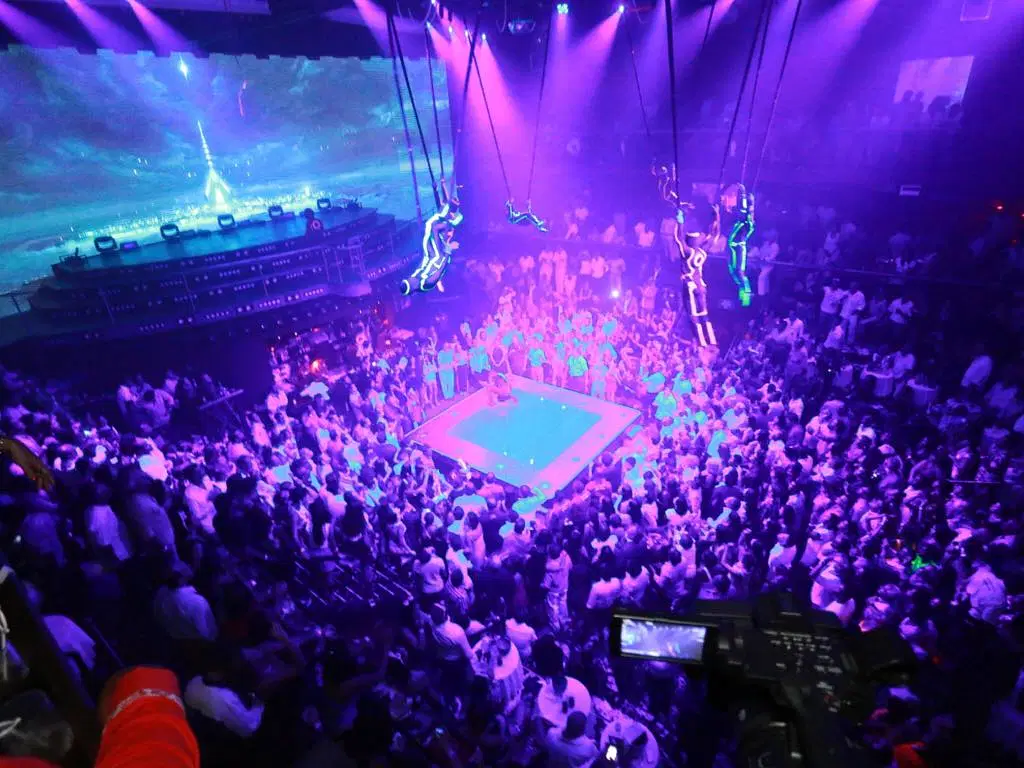 A large crowd surrounds a blue-lit boxing ring in a nightclub with vivid purple lighting—an electrifying scene to experience on your punta cana trip, where performers in glowing suits and digital screens create a futuristic atmosphere.