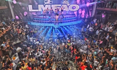 A crowded nightclub in Punta Cana pulses with energy as people dance and cheer around a large stage. Performers, bright lights, confetti, and an “LMFAO” sign create a festive vibe—a must-see spot during Punta Cana excursions.