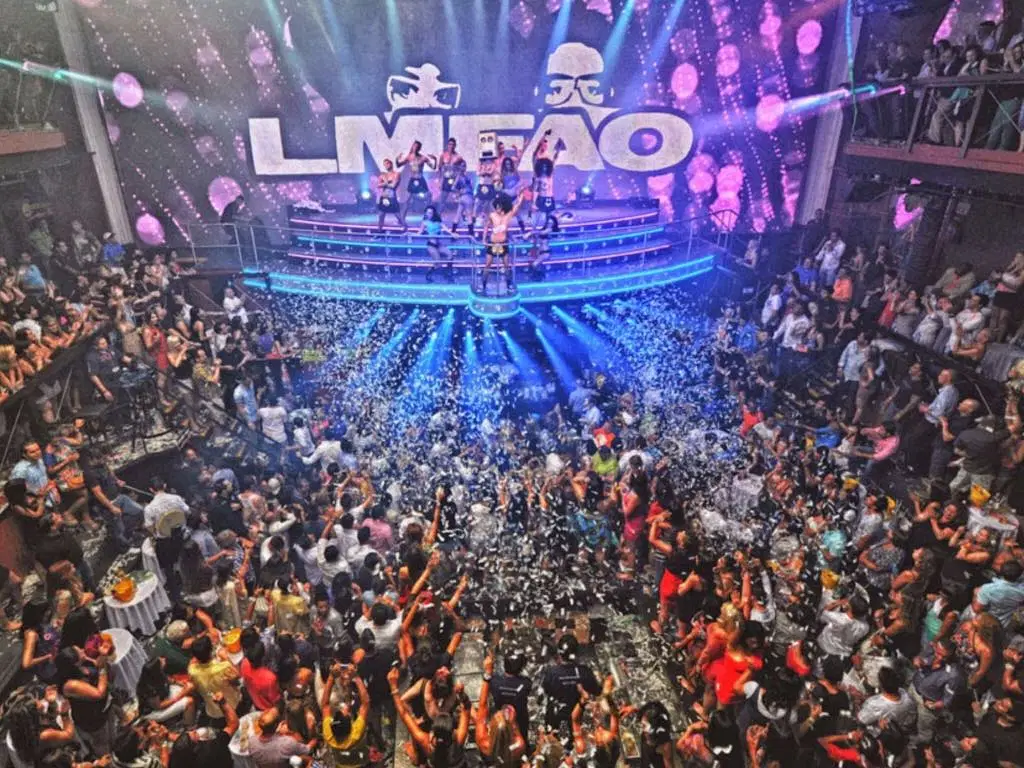 A crowded nightclub in Punta Cana pulses with energy as people dance and cheer around a large stage. Performers, bright lights, confetti, and an “LMFAO” sign create a festive vibe—a must-see spot during Punta Cana excursions.
