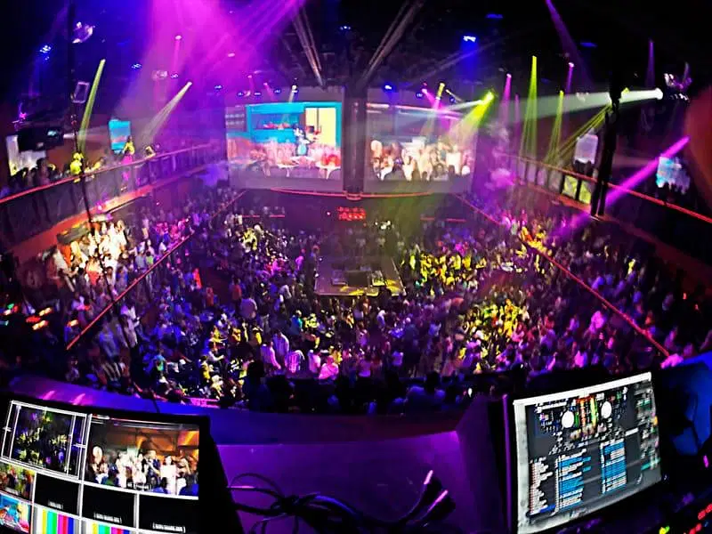A crowded nightclub on your Punta Cana trip, filled with people dancing under vibrant neon lights. The photo, taken from the DJ booth, overlooks the energetic dance floor and two upper balconies—an unforgettable highlight among Punta Cana excursions.