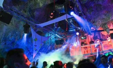 A crowded underground nightclub in Punta Cana, where people dance under colorful lights and fog. The rocky cave ceiling looms above as laser beams flash and a neon-lit DJ booth sets the scene—one of the most exciting Punta Cana excursions for nightlife lovers.