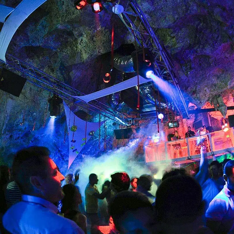 A crowded underground nightclub in Punta Cana, where people dance under colorful lights and fog. The rocky cave ceiling looms above as laser beams flash and a neon-lit DJ booth sets the scene—one of the most exciting Punta Cana excursions for nightlife lovers.