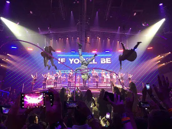 A vibrant stage show, part of exciting tours in Punta Cana, features acrobats suspended mid-air under colorful lights. A lively crowd captures the moment as a band and singers perform on multi-tiered platforms beneath a large lit-up sign.