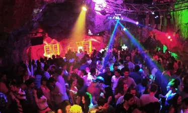 A large crowd dances in a vibrant, rocky cave nightclub filled with colorful lights—purple, blue, green, and yellow. This lively hotspot is a must-see on popular Punta Cana tours for those craving energy and excitement.