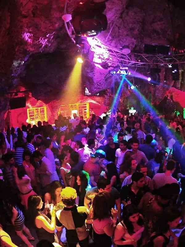A large crowd dances in a vibrant, rocky cave nightclub filled with colorful lights—purple, blue, green, and yellow. This lively hotspot is a must-see on popular Punta Cana tours for those craving energy and excitement.