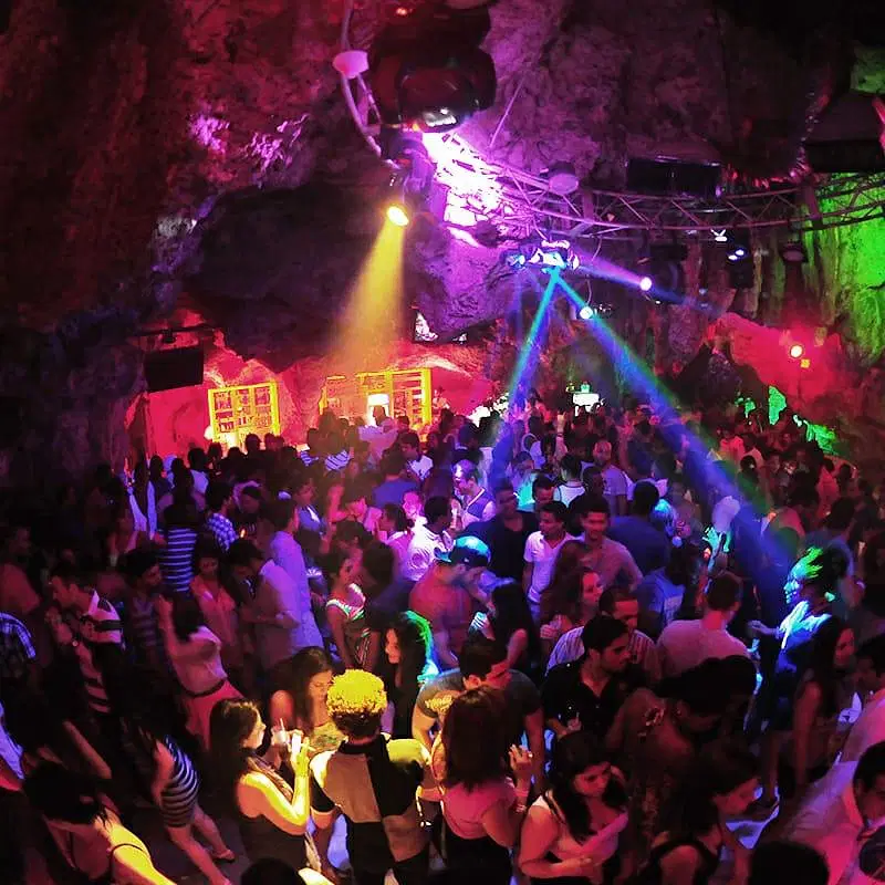 A large crowd dances in a vibrant, rocky cave nightclub filled with colorful lights—purple, blue, green, and yellow. This lively hotspot is a must-see on popular Punta Cana tours for those craving energy and excitement.