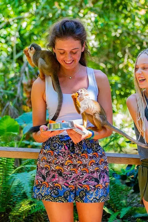 A smiling woman in a white tank top and patterned shorts holds a tray with food as two small monkeys eat from it, one perched on her shoulder. Lush green foliage fills the background—a fun moment during tours in Punta Cana.