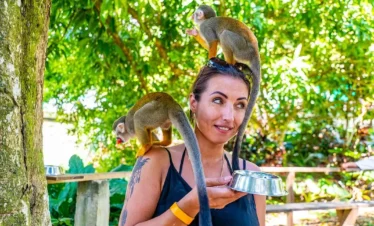 A woman outdoors holds a metal bowl in one hand while two small monkeys climb on her, one perched on her head and the other on her shoulder. She smiles and looks to the side—capturing the fun of Punta Cana tours amid green trees and sunlight.