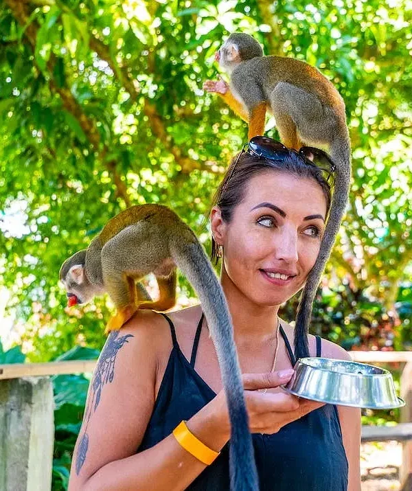 A woman outdoors holds a metal bowl in one hand while two small monkeys climb on her, one perched on her head and the other on her shoulder. She smiles and looks to the side—capturing the fun of Punta Cana tours amid green trees and sunlight.