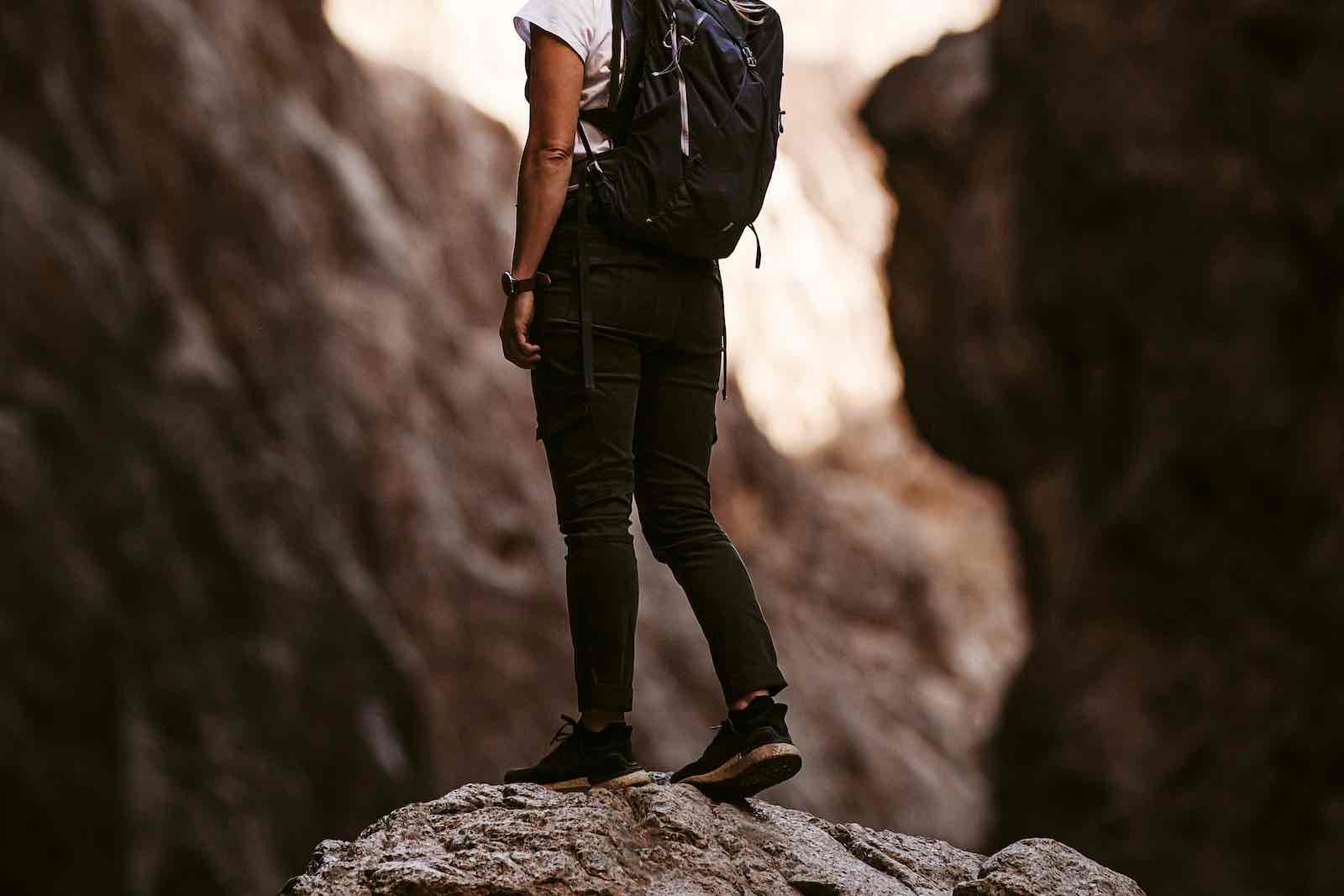 A person stands alone on a large rock in a rugged canyon, wearing a backpack, black pants, and sneakers. Sunlight highlights the canyon walls—a scene that emphasizes solo travel safety in adventurous landscapes.