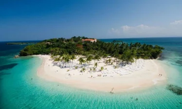 Aerial view of a small tropical island with white sandy beaches, turquoise water, and palm trees. Ideal for Punta Cana excursions, the island features beach chairs and umbrellas, lush greenery at its center, and a sunny blue sky overhead.