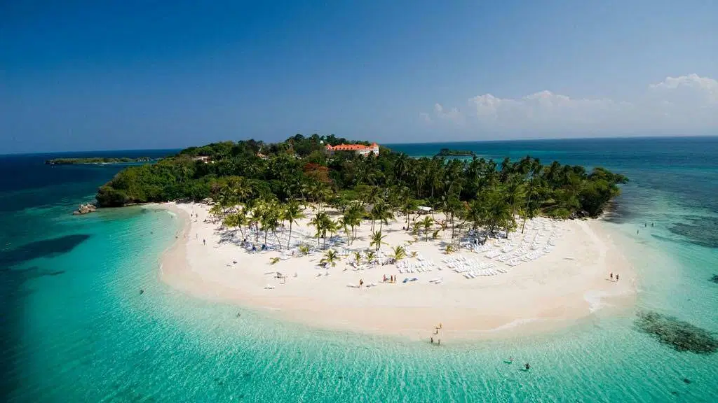 Aerial view of a small tropical island with white sandy beaches, turquoise water, and palm trees. Ideal for Punta Cana excursions, the island features beach chairs and umbrellas, lush greenery at its center, and a sunny blue sky overhead.