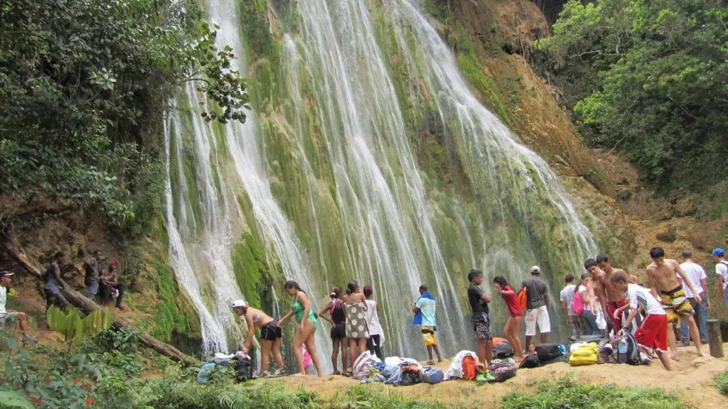 A group in swimwear gathers at the base of a lush, moss-covered waterfall, surrounded by vibrant vegetation—some stand while others relax near their bags and towels, savoring a day in nature on one of many unforgettable Punta Cana excursions.
